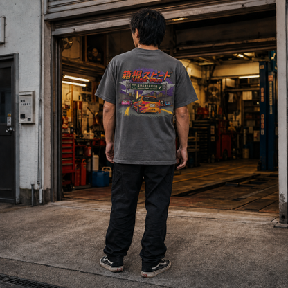 A man stands with his back to the camera in front of an open garage door, wearing a dark gray t-shirt with a colorful Japanese graphic print on the back, black pants, and black sneakers.