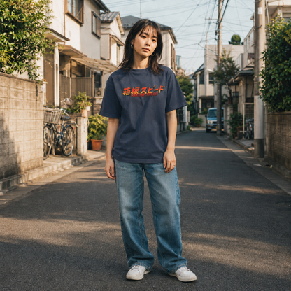 A woman stands on a sunlit residential street wearing a navy oversized graphic T-shirt with red and orange Japanese text across the chest, light blue wide-leg jeans, and white sneakers.