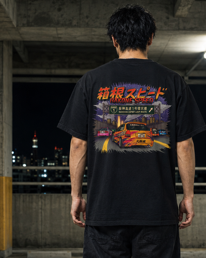 A man stands with his back to the camera in a concrete parking garage at night, wearing a black graphic T-shirt and dark pants. The shirt features a large, colorful Japanese street racing graphic on the back with a red car and stylized text.